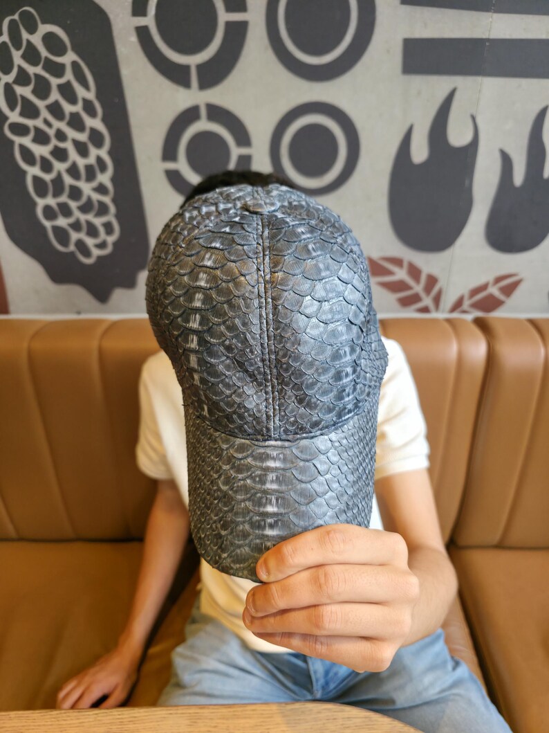 May include: A dark gray baseball cap with a textured, scaly pattern. The cap has a curved brim and a structured crown. The material appears to be leather or a leather-like material. The cap is being held up in front of a person.