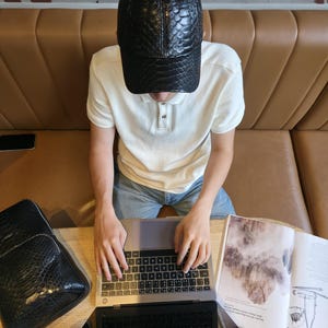 K&ouml;nnte beinhalten: Schwarze Baseballkappe aus Leder mit Schlangenmuster. Eine Person tr&auml;gt die Kappe und arbeitet an einem Laptop. Ein passender schwarzer Rucksack ist ebenfalls zu sehen. Die Szene spielt in einem Caf&eacute;.