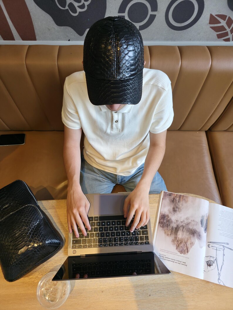 K&ouml;nnte beinhalten: Schwarze Baseballkappe aus Leder mit Schlangenmuster. Eine Person tr&auml;gt die Kappe und arbeitet an einem Laptop. Ein passender schwarzer Rucksack ist ebenfalls zu sehen. Die Szene spielt in einem Caf&eacute;.
