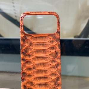 May include: Orange and brown snakeskin-textured phone case. The case has a rectangular shape with rounded corners and a camera cutout. The case is made of a material that mimics the look of snakeskin.