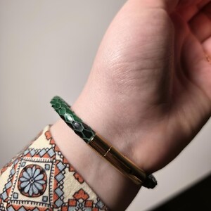 以下が含まれることがあります： 緑と黒のヘビ柄のブレスレット、ゴールドカラーの留め金付き。 ブレスレットは手首に着用され、背景には模様入りの袖が見えます。