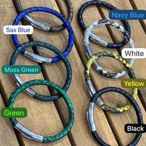 May include: A collection of braided leather bracelets in various colors, including brown, navy blue, white, yellow, and black. Each bracelet features a silver-colored magnetic clasp. The bracelets are displayed on a wooden surface.