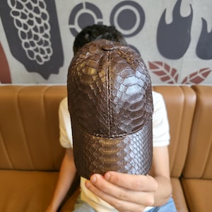 Peut inclure: Casquette de baseball marron avec un motif peau de serpent. La casquette a une visière incurvée et une couronne structurée. La casquette est tenue par une personne. La casquette est faite d'un matériau texturé.