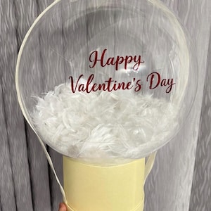 May include: A clear, round balloon with the text "Happy Valentine's Day" in red script. The balloon is filled with white feathers and sits atop a pale yellow cylindrical container. A white ribbon is tied at the top.