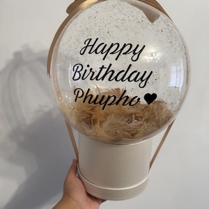 Puede incluir: Un globo redondo transparente con el texto "Happy Birthday Phupho" en escritura negra, lleno de purpurina dorada y plumas marrones. El globo se asienta sobre una caja cilíndrica color crema con un asa de cinta dorada.