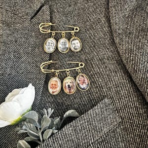 以下が含まれることがあります： 小さな写真が収められた3つの楕円形のペンダントが付いた、2つの金色の安全ピン。 ピンは、ジャケットまたはコートと思われる、質感のある灰色の生地にセットされています。 白い花と緑の葉が左下隅にあります。