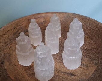 May include: Seven clear, tower-shaped selenite crystals are displayed in a wooden bowl. The crystals have a layered appearance and are translucent. The bowl is made of wood and has a natural grain.