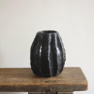Könnte beinhalten: Eine schwarze, strukturierte Vase mit vertikalen Rippen. Die Vase hat eine abgerundete Form und steht auf einer Holzoberfläche. Die dunkle Farbe und die einzigartige Textur verleihen ihr ein rustikales, handwerkliches Aussehen. Diese dekorative Vase eignet sich für die Inneneinrichtung.