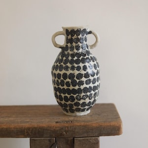 Może przedstawiać: Ceramiczny wazon z dwoma uchwytami, zdobiony wzorem czarnych kropek na kremowym tle. Wazon stoi na rustykalnej drewnianej ławce. Wazon ma zaokrąglony kształt i lekko teksturowaną powierzchnię.