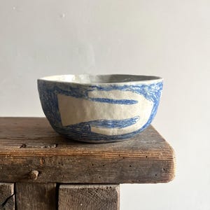Peut inclure: Un bol en céramique avec une base blanc cassé et un motif bleu texturé dessiné à la main, composé de formes et de lignes abstraites. Le bol présente un bord irrégulier et organique ainsi qu'une surface légèrement inégale.