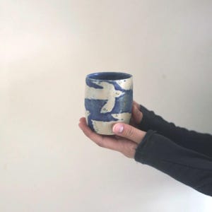 Peut inclure: Une tasse cylindrique en céramique avec un intérieur bleu et un extérieur crème. La tasse présente un motif répétitif de figures blanches stylisées sur un fond bleu. La tasse est tenue dans deux mains.