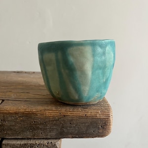 May include: A small, hand-thrown ceramic cup with a rounded, organic shape. The exterior features a reactive glaze with vertical streaks of vibrant turquoise and muted cream, creating a textured, layered appearance with a visible, uneven rim.