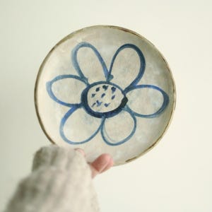 Könnte beinhalten: Ein kleiner, runder Keramikteller mit einem handgemalten blauen Blumenmuster. Die Blume hat sechs Blütenblätter und eine dunkelblaue Mitte. Der Teller hat einen cremefarbenen Hintergrund und einen braunen Rand. Der Teller wird von einer Person gehalten.