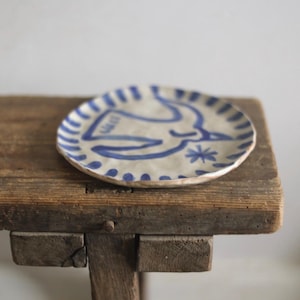 Op de afbeelding: Een ronde keramische plaat met een handgeschilderd blauw vogelontwerp. De plaat heeft een witte achtergrond met een blauwe rand en rust op een rustiek houten oppervlak. De vogel is afgebeeld met eenvoudige lijnen en een ster.