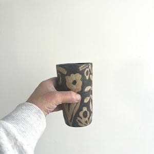 Peut inclure: Un gobelet cylindrique en céramique présentant une base de couleur noir charbon foncé avec un motif floral beige clair contrastant sculpté dans la surface. Le récipient a une texture mate et un aspect artisanal aux formes organiques.