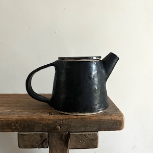 May include: A black ceramic teapot with a glossy, uneven surface texture, a wide handle, a short spout, and a flat lid. The teapot features a slightly flared base and a dark, reflective finish that highlights the organic, hand-formed shape of the vessel.