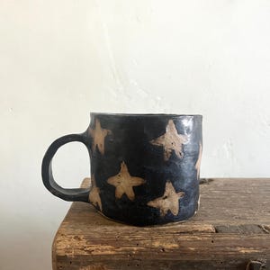 Peut inclure: Une tasse en céramique faite à la main avec un vernis noir foncé et une série de formes d'étoiles à cinq branches beige clair réparties sur la surface extérieure. La tasse présente une anse arrondie et une forme organique légèrement irrégulière.