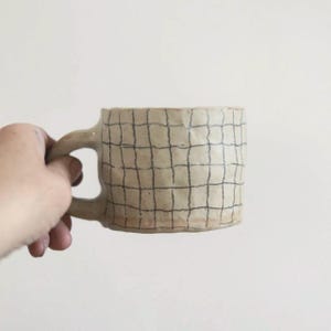 Może przedstawiać: Ręcznie wykonany ceramiczny kubek z kremową podstawą i wzorem kratki w kolorze ciemnoniebieskim. Kubek ma kwadratowy kształt i zaokrąglony uchwyt. Kubek jest trzymany przez dłoń na białym tle.