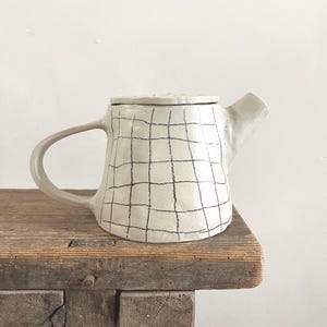 Può includere: Una teiera in ceramica fatta a mano con smalto bianco sporco e un motivo a griglia nero disegnato a mano. La teiera presenta un'ampia impugnatura, un beccuccio corto e un coperchio piatto abbinato, con una forma organica e irregolare.
