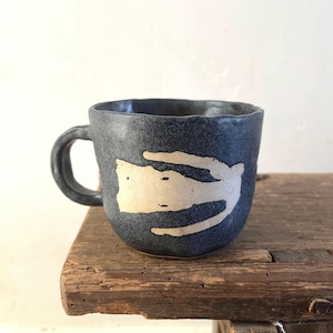 May include: A dark grey, speckled ceramic mug with a hand-formed, irregular rim and a curved handle. The exterior features a prominent, light beige, abstract organic shape painted across the centre, contrasting with the dark, textured surface.