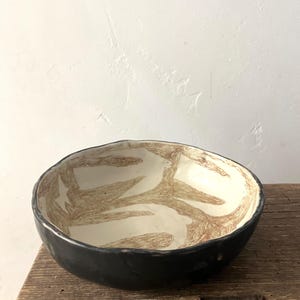 Op de afbeelding: Een ronde, ondiepe keramische kom met een onregelmatige, organische rand. De buitenkant is effen matzwart, terwijl de binnenkant een crèmekleurige basis heeft met een bruin, getextureerd, abstract patroon dat doet denken aan takken.