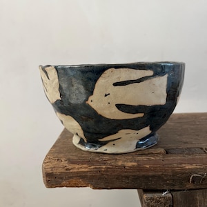 Peut inclure: Un bol en céramique avec une glaçure bleu foncé brillante présentant un motif répétitif de silhouettes d'oiseaux stylisées de couleur crème. Le bol a un bord irrégulier formé à la main et une petite base circulaire surélevée, avec une finition texturée.