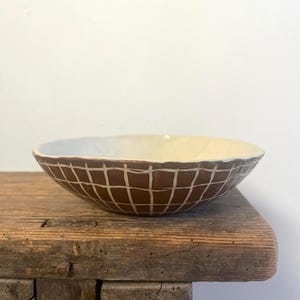 Pode incluir: Uma tigela de cerâmica rasa com um exterior castanho escuro apresentando um padrão de grelha branco. O interior da tigela é de uma cor esbranquiçada lisa. A borda da tigela tem uma forma irregular feita à mão com um perfil ligeiramente ondulado.
