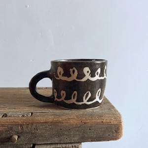 Può includere: Una tazza in ceramica marrone scuro con un manico arrotondato e un corpo cilindrico largo. La superficie esterna presenta due file orizzontali di motivi a cappio continui dipinti a mano in bianco sporco che circondano la tazza.