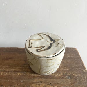 以下が含まれることがあります： クリーム色のセラミックジャー、蓋付き。 蓋には茶色の鳥のデザインがあります。 ジャーの本体は、茶色の星と線の模様で装飾されています。 ジャーは木製の表面に置かれています。
