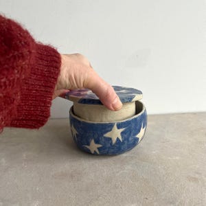 Peut inclure: Un bol en céramique bleu avec un couvercle, décoré d'étoiles blanches. Le bol est rond et possède un couvercle assorti. Le bol est ouvert par une main. Le bol est probablement utilisé pour le stockage ou l'affichage.