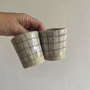 以下が含まれることがあります： 手描きの黒い格子模様が施されたオフホワイトの陶器製カップ2個。カップはわずかに不規則で有機的な形状をしており、上部は光沢のある仕上げ、底面はマットで釉薬のかかっていない質感になっています。シンプルな正方形のグリッドデザインです。