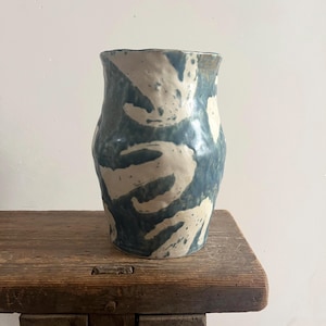 Peut inclure: Un vase en céramique avec une silhouette façonnée à la main, légèrement irrégulière. La surface présente un motif abstrait audacieux de coups de pinceau bleu-gris profond sur une base blanc cassé, créant un aspect visuel texturé et organique.