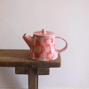 Peut inclure: Une théière en céramique rose clair ornée d'un motif peint à la main de fleurs rouges et orange. La théière possède un couvercle assorti avec un petit bouton, un bec verseur incurvé et une anse latérale, le tout avec une finition émaillée.