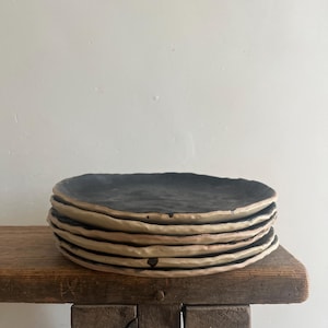 Pode incluir: Uma pilha de cinco pratos circulares com uma superfície interior preto mate e uma borda exterior bege claro contrastante. Cada prato apresenta uma borda irregular formada à mão com um acabamento texturizado, criando um aspeto em camadas.