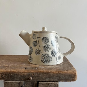 Op de afbeelding: Een crèmekleurige keramische theepot met een handgetekende zwarte schets van een tak met bladeren die rondom de pot loopt. De theepot heeft een brede basis, een korte tuit, een gebogen handvat en een bijpassend deksel met een kleine knop.