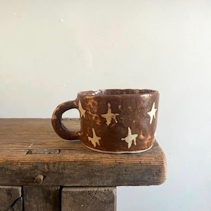 May include: A brown ceramic mug with a glossy finish and a hand-formed, irregular shape. The exterior surface is decorated with multiple white, hand-painted star motifs scattered across the body. It features a thick, rounded handle on the side.