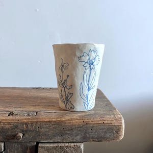 Könnte beinhalten: Ein weißer Keramikbecher mit unebener, strukturierter Oberfläche, der mit handgezeichneten blauen botanischen Illustrationen von zwei verschiedenen Blumen verziert ist. Der Becher hat einen leicht ausgestellten Rand und einen konischen Boden.
