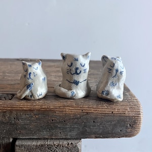 May include: Three small, hand-sculpted ceramic cat figurines in a light off-white glaze. Each cat features simple, hand-drawn blue ink details depicting facial expressions, ears, and tails. The figures are arranged in a row, showcasing varied poses.