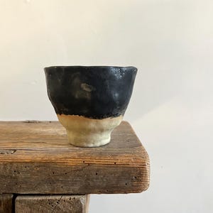 Könnte beinhalten: Eine kleine, handgefertigte Keramiktasse mit zweifarbigem Design. Der obere Teil der Tasse ist mit einer mattschwarzen Glasur versehen, während der untere Sockel aus unglasiertem, strukturiertem beigefarbenem Ton mit unregelmäßigem Rand besteht.