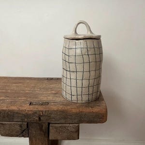 Op de afbeelding: Een cilindrische keramische pot met een gespikkeld gebroken wit glazuur en een handgetekend zwart ruitpatroon. De pot heeft een bijpassend deksel met een kleine, boogvormige handgreep aan de bovenkant en een getextureerde, organische afwerking.