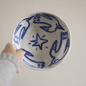 Op de afbeelding: Een witte keramische kom met een blauw handgeschilderd ontwerp van vogels en een ster. De kom wordt door een persoon vastgehouden, waarbij het interieurkunstwerk wordt getoond. Het ontwerp is eenvoudig en modern, geschikt voor het serveren van voedsel of als decoratief stuk.