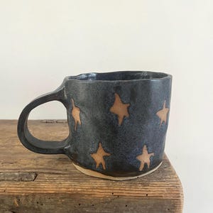 Op de afbeelding: Een handgemaakte keramische mok met een donkerblauw, gespikkeld glazuur en een reeks lichtbruine, vierpuntige stervormen verspreid over het buitenoppervlak. De mok heeft een breed, gebogen handvat en een onregelmatige, organische randvorm.