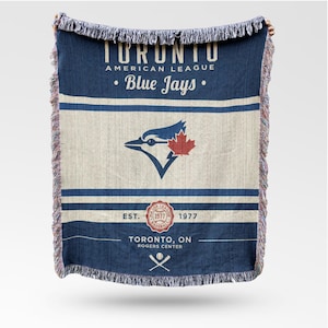 Op de afbeelding: Een blauw en crèmekleurig geweven sprei met de tekst "TORONTO AMERICAN LEAGUE Blue Jays." De sprei heeft een logo van een blauwe gaai met een rood esdoornblad. De onderkant van de sprei heeft de tekst "EST. 1977 TORONTO, ON ROGERS CENTER."