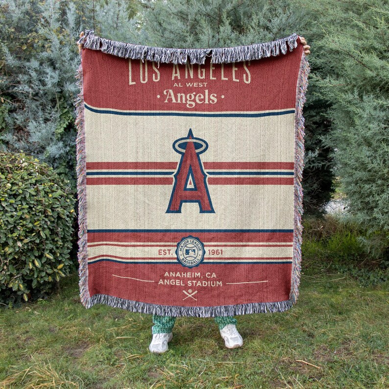 Puede incluir: Una manta tejida roja, beige y azul con el texto "LOS ANGELES AL WEST Angels" y el logotipo del equipo. La manta tambi&eacute;n incluye el texto "EST. 1961" y "ANAHEIM, CA ANGEL STADIUM". La manta tiene un borde con flecos.