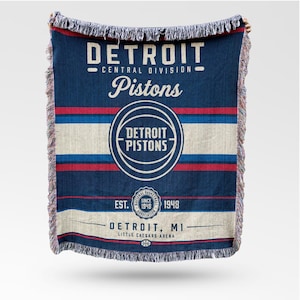 Op de afbeelding: Een blauw-rood gestreepte deken met de tekst "DETROIT Pistons" en het teamlogo. De deken bevat de tekst "Central Division", "EST. 1948" en "Little Caesars Arena". De deken heeft een gefranjerde rand.