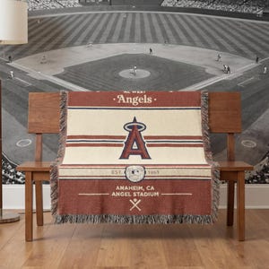 Puede incluir: Una manta tejida con el logotipo de los Angelinos de Los &Aacute;ngeles en rojo, blanco y azul marino. La manta muestra el nombre del equipo, el logotipo y la informaci&oacute;n del estadio. La manta tiene un borde con flecos y est&aacute; colocada sobre un banco de madera.