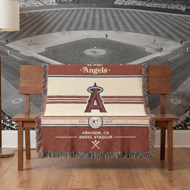 Puede incluir: Una manta tejida con el logotipo de los Angelinos de Los &Aacute;ngeles en rojo, blanco y azul marino. La manta muestra el nombre del equipo, el logotipo y la informaci&oacute;n del estadio. La manta tiene un borde con flecos y est&aacute; colocada sobre un banco de madera.