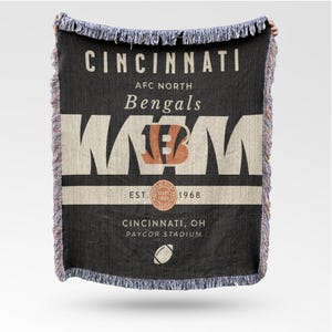 Può includere: Coperta nera tessuta con testo bianco "CINCINNATI AFC NORTH Bengals". La coperta presenta il logo della squadra e "EST. 1968". La parte inferiore della coperta recita "CINCINNATI, OH PAYCOR STADIUM" con una grafica di calcio. I bordi hanno un bordo a frange.