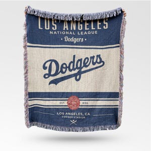 Op de afbeelding: Een blauw en beige geweven sprei met de tekst "LOS ANGELES NATIONAL LEAGUE • Dodgers." Het woord "Dodgers" staat in een scriptlettertype. De sprei bevat ook de tekst "EST. 1890" en "LOS ANGELES, CA DODGER STADIUM."