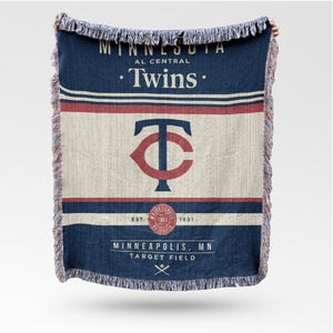 Puede incluir: Una manta azul marino y crema con el logotipo y el texto de los Minnesota Twins. La manta tiene un borde con flecos e incluye las palabras "Twins", "Minneapolis, MN" y "Target Field".